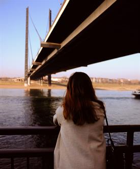 a young woman with a bright coat and brown hair is standing at a banister and watching over a big river. A dark bridge is obove her.